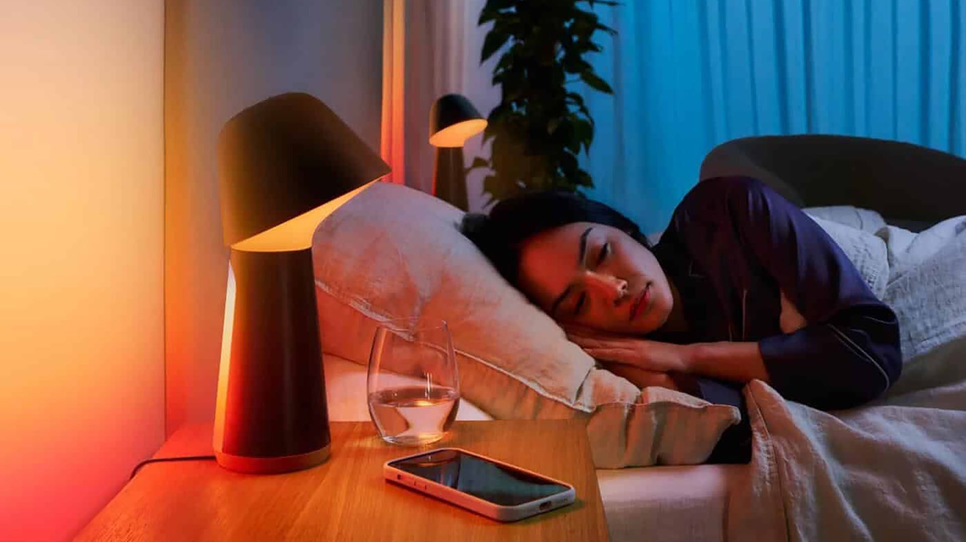 A woman sleeping in a bed next to a smart lamp and a smartphone.