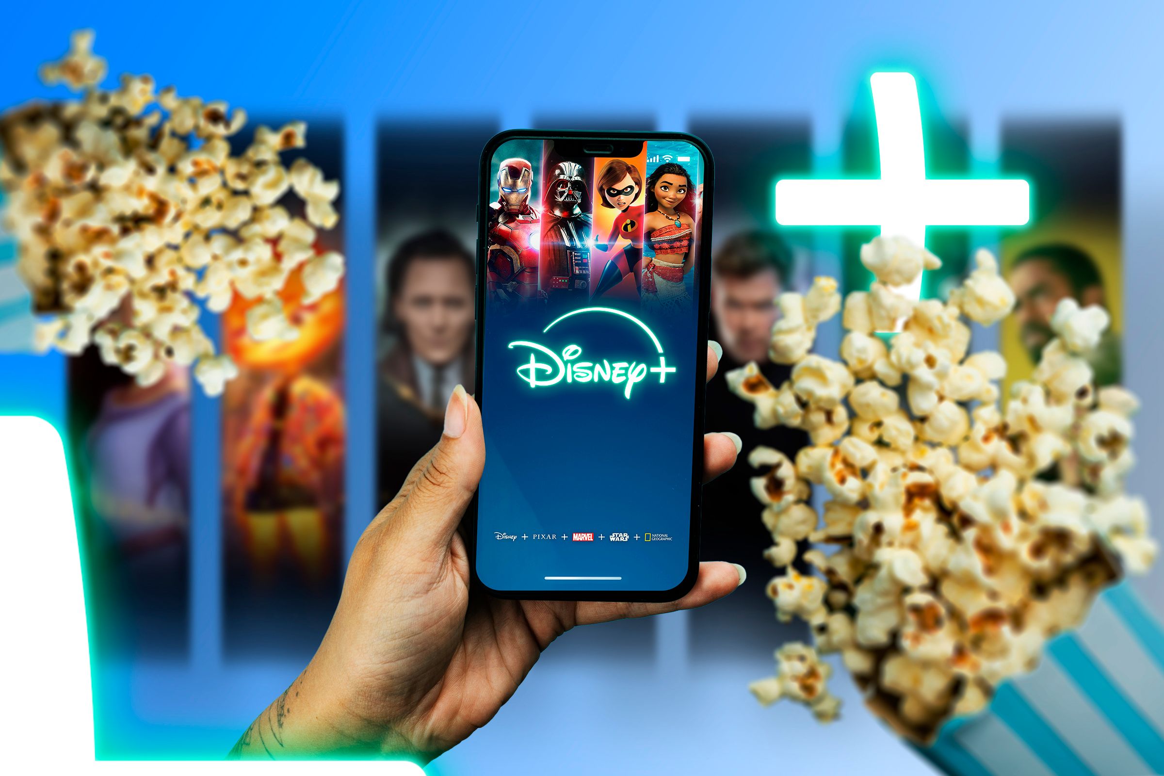 A hand holding a phone with the Disney+ screen displayed, and two popcorn bags next to it.