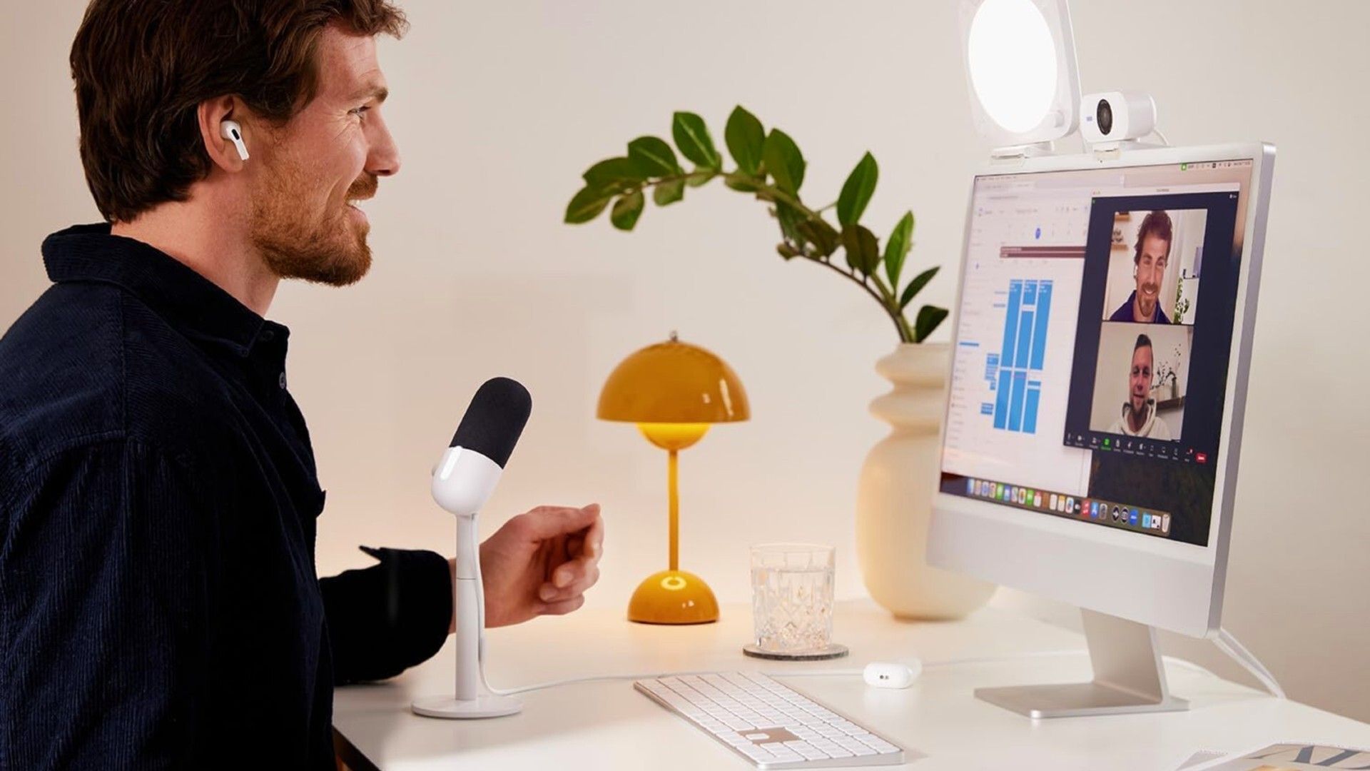 A man uses a computer for a video call, with a microphone and lamp on his desk, and a plant in the background.