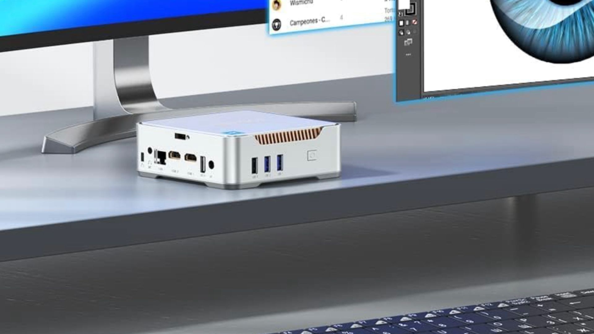 A Mini PC sitting on a desk under computer monitors.